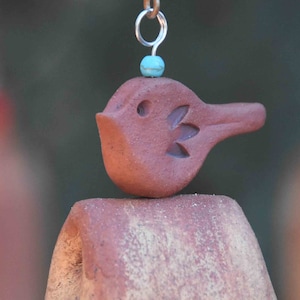 May include: A terracotta-colored clay bird ornament with a carved wing detail, featuring a small turquoise bead positioned above its head and a silver-toned metal jump ring attached to the top for hanging purposes, resting on a curved clay base.