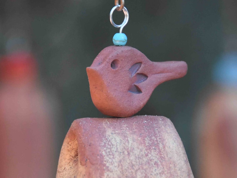 May include: A terracotta-colored clay bird ornament with a carved wing detail, featuring a small turquoise bead positioned above its head and a silver-toned metal jump ring attached to the top for hanging purposes, resting on a curved clay base.