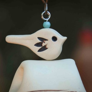 May include: A cream-colored ceramic bird ornament with a small dark eye and three dark leaf-shaped markings on its side. The bird is topped with a light blue bead and a silver metal jump ring, suspended from a thin brown cord against a dark background.