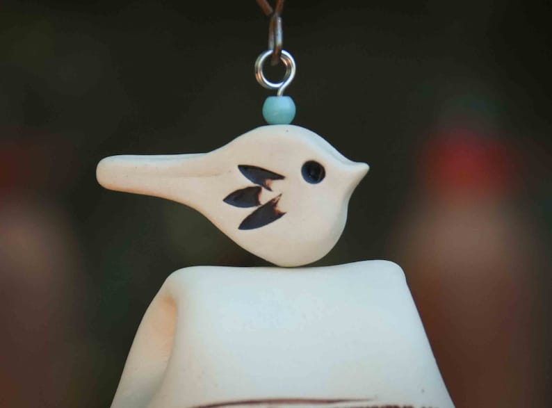 May include: A cream-colored ceramic bird ornament with a small dark eye and three dark leaf-shaped markings on its side. The bird is topped with a light blue bead and a silver metal jump ring, suspended from a thin brown cord against a dark background.