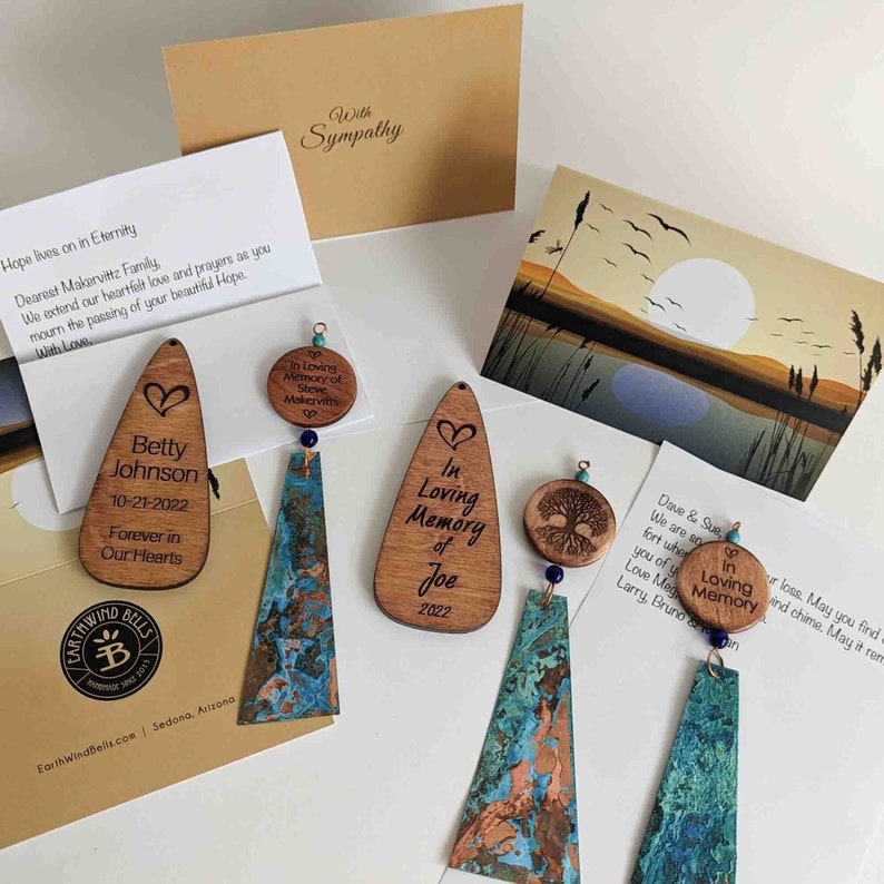 May include: Memorial wind chimes featuring wooden teardrop and circular plaques engraved with names, dates, and tree motifs. The chimes include textured, oxidized copper-toned metal panels, small blue beads, and accompanying sympathy cards with printed messages.