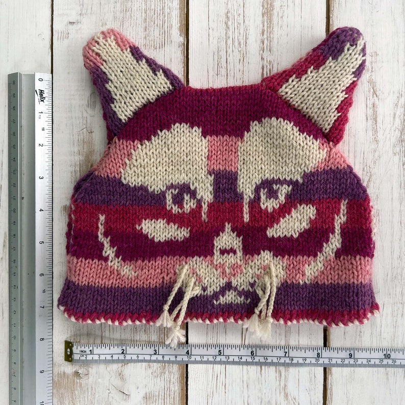 a pink striped knitted cat tea cosy with rulers and tape measure to show size