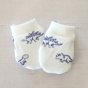 May include: A pair of white baby mittens with a blue dinosaur pattern. The mittens are made of soft fabric and feature a rolled cuff. The dinosaur illustrations include a T-Rex and Stegosaurus.