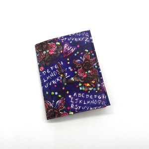May include: A purple wallet with a Stranger Things theme. The wallet features a graphic of the Demogorgon, Christmas lights, and the alphabet written in white. The text "Run" is written in white on the wallet.