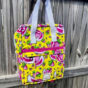 May include: A yellow and pink tote bag with a strawberry print and white straps. The bag has a small patch with the text "The Wild Wife" on it.