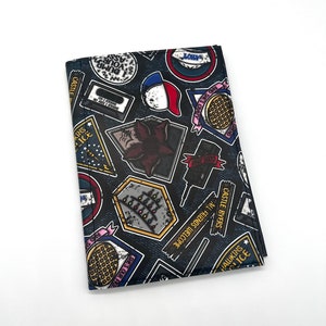May include: A black wallet with a denim-like texture and a variety of colorful patches featuring text and images from the show Stranger Things. The patches include the words "Hawkins", "Castle Byers", "All Friends Welcome", "She's Our Friend", "Should I Go", "Dump Your Ass", "11", "L.O.V.E", "And See What Happens", "Dress Our Friends", "Modern Soreva TV", "Castle Byers", "Hawkins Police", and "1983".