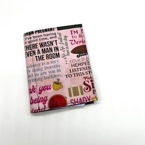 May include: Pink fabric wallet with a black border. The wallet is covered in a pattern of text and images, including the text "I'm to be Verd" and "Shady".