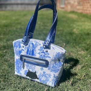 May include: A white handbag with a blue and white floral pattern featuring a Star Wars theme. The bag has blue handles and a zipper closure.