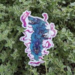Puede incluir: Una pegatina con un diseño de lobo de tres cabezas en tonos azules y morados. Las cabezas de lobo tienen múltiples ojos y dientes afilados, rodeadas de remolinos morados en forma de humo. La pegatina está sobre un fondo de follaje verde.