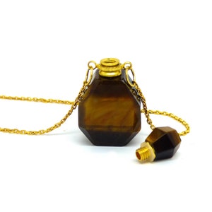 Puede incluir: Un collar de cadena dorada con un colgante de piedra de ojo de tigre facetado. El colgante tiene forma de botella con una tapa dorada y un tap&oacute;n separado de piedra de ojo de tigre.