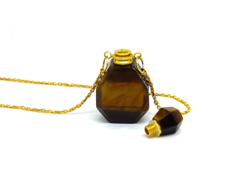 Puede incluir: Un collar de cadena dorada con un colgante de piedra de ojo de tigre facetado. El colgante tiene forma de botella con una tapa dorada y un tap&oacute;n separado de piedra de ojo de tigre.
