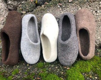 Puede incluir: Cinco pares de zapatillas de fieltro en diferentes colores: marrón, gris, blanco, gris oscuro y marrón claro. Las zapatillas están alineadas en una superficie cubierta de musgo.