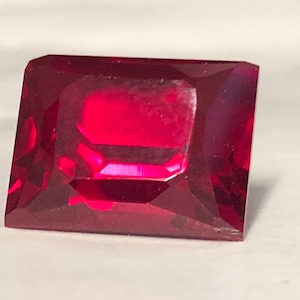 May include: A rectangular faceted gemstone with a deep, vibrant red color. The stone features a clear, polished surface with multiple geometric facets that reflect light, creating sharp edges and a brilliant, transparent appearance throughout.