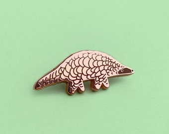 May include: A gold-coloured enamel pin featuring a pangolin, a small mammal with scales, in a walking pose. The pin is on a green background.