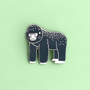 Peut inclure: Épingle à nourrice représentant un gorille gris foncé avec des détails blancs et un contour blanc. Le gorille a une expression sérieuse. L'épingle est posée sur un fond vert clair.