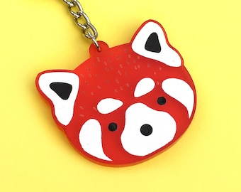 May include: Red panda keychain with a white face and black ears. The keychain is made of red plastic and has a silver metal ring.