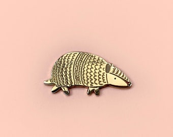 May include: Gold-coloured armadillo pin with a detailed design. The armadillo is facing right and has a textured shell.