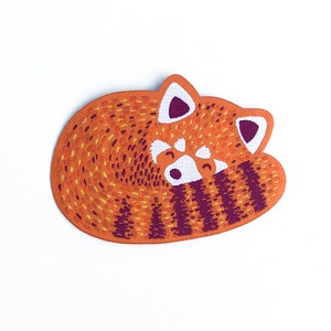 May include: An orange and brown patch featuring a stylized red panda curled up in a sleeping position. The patch has white accents on the face and ears, and dark purple stripes. The patch is on a white background.
