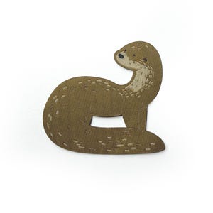 May include: A brown embroidered patch in the shape of an otter. The otter is depicted in a seated position with its body curved, featuring a lighter tan face, a small black nose, and subtle stitched details throughout its textured brown body.