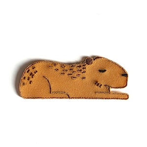 Peut inclure: Un écusson brodé de couleur beige représentant un capybara, un grand rongeur d'Amérique du Sud, en position couchée. L'écusson a des yeux noirs et de petites taches brunes. Fond blanc.