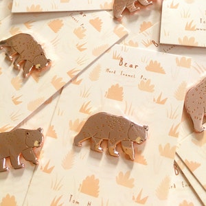 Può includere: Spille a forma di orso in smalto color oro rosa su biglietti con un design arancione chiaro e beige. Le spille hanno la forma di un orso che cammina. Le carte presentano il testo "Bear Hard Enamel Pin" e il sito web "www.tom-hardwick.uk".