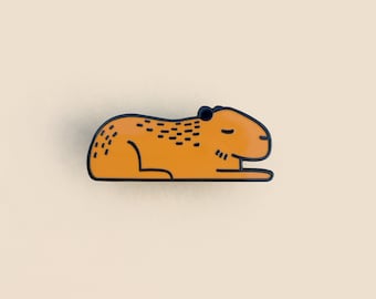 May include: Orange enamel pin of a capybara lying down with its eyes closed. The pin has a black outline and a dotted pattern on its back.