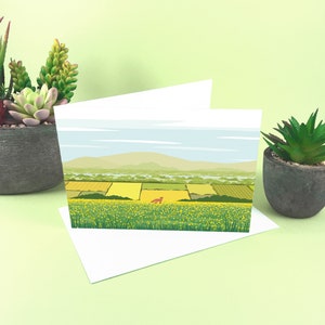 May include: A blank greeting card with a landscape scene of a field of yellow wildflowers and a small red fox in the distance. The card is folded in half and has a white envelope.