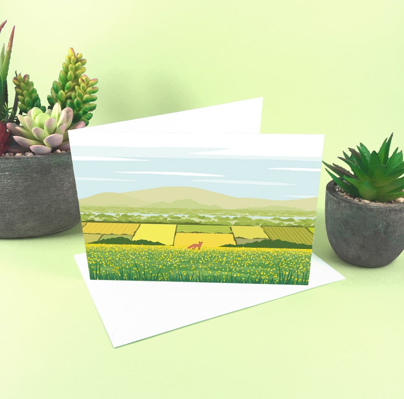 May include: A blank greeting card with a landscape scene of a field of yellow wildflowers and a small red fox in the distance. The card is folded in half and has a white envelope.