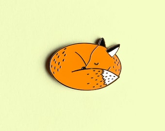 May include: A gold-colored enamel pin featuring a curled-up orange fox with a black nose and eyes. The fox is sleeping with its tail curled around its body.