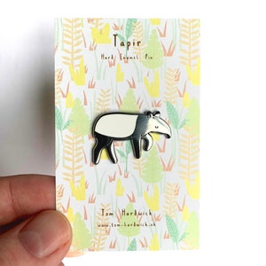 May include: A silver enamel pin featuring a cartoon tapir with a white stripe on its back. The pin is on a card with a jungle-themed background and the text "Tapir Hard Enamel Pin" and "Tom Hardwick www.tom-hardwick.uk".