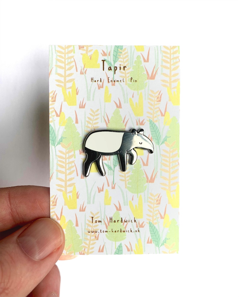 May include: A silver enamel pin featuring a cartoon tapir with a white stripe on its back. The pin is on a card with a jungle-themed background and the text "Tapir Hard Enamel Pin" and "Tom Hardwick www.tom-hardwick.uk".