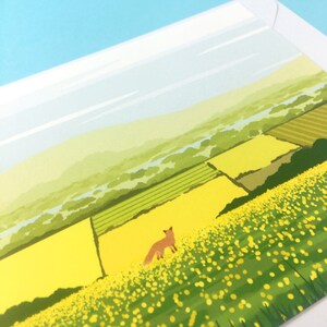 May include: A postcard featuring a stylized illustration of a fox standing in a field of yellow wildflowers. The background shows a rolling green landscape with a river winding through it.
