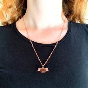 May include: A rose gold necklace featuring a small axolotl charm. The axolotl is pink with black eyes and a small smile.