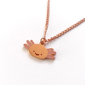 May include: A rose gold necklace featuring a small axolotl charm. The axolotl is a cartoon-style illustration with a pink body and a happy face.
