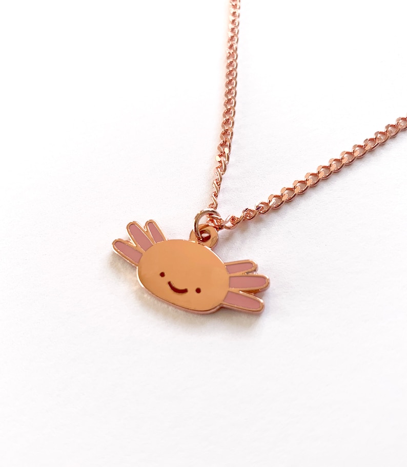 May include: A rose gold necklace featuring a small axolotl charm. The axolotl is a cartoon-style illustration with a pink body and a happy face.