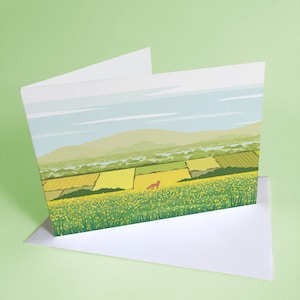 May include: A greeting card with a landscape scene of a field of yellow wildflowers with a red fox in the distance. The sky is blue with white clouds and there are green hills in the background.