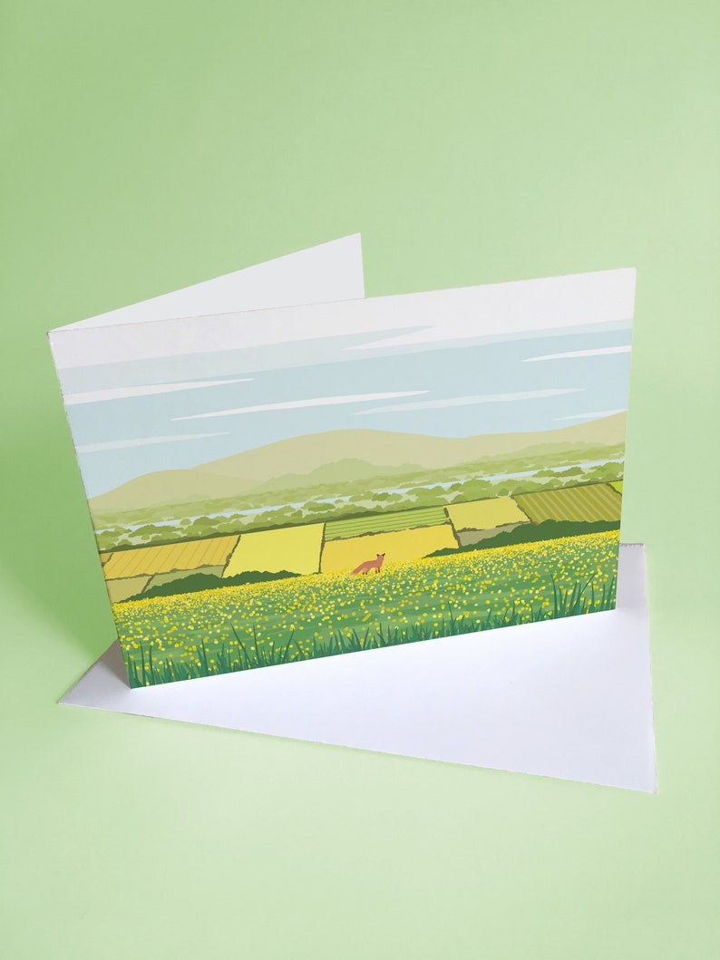 May include: A greeting card with a landscape scene of a field of yellow wildflowers with a red fox in the distance. The sky is blue with white clouds and there are green hills in the background.