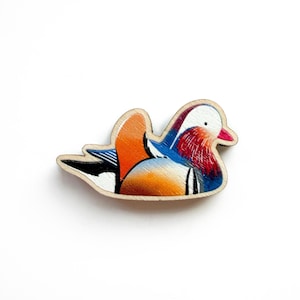 Op de afbeelding: Houten speld met een ontwerp van een mandarijneend. De eend heeft een witte kop, een rode borst, blauwe vleugels en oranje en bruine accenten. De speld is in de vorm van de eend uitgesneden en staat tegen een witte achtergrond.
