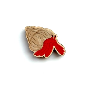 May include: Wooden hermit crab pin with a red body and a tan shell. The crab is painted with a detailed design, and the pin is set against a white background. This decorative pin is perfect for adding a touch of coastal charm to any outfit.