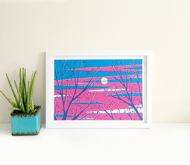 May include: A framed art print featuring a stylized landscape with a pink and blue sky, white clouds, and silhouetted trees. The artwork has a modern, graphic style. A small, turquoise square planter with a green plant is to the left of the framed print.