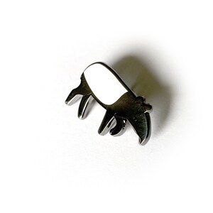 May include: A silver-colored metal pin shaped like a tapir. The tapir is facing left and has a white body and a black head and snout.
