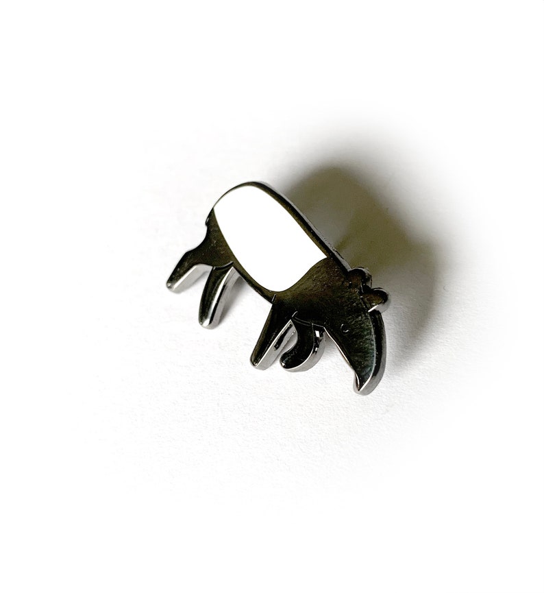 May include: A silver-colored metal pin shaped like a tapir. The tapir is facing left and has a white body and a black head and snout.