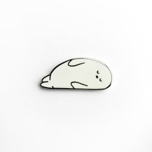 Op de afbeelding: Witte emaille pin met een zwarte omtrek van een cartoon zeehond die op zijn rug ligt met zijn ogen dicht.