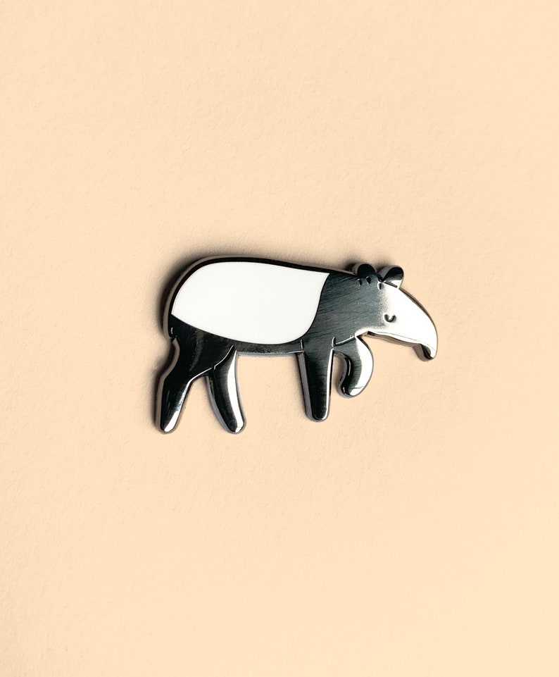 May include: A silver-colored enamel pin featuring a cartoon depiction of a tapir. The tapir is white with a black snout and legs. The pin has a shiny finish.