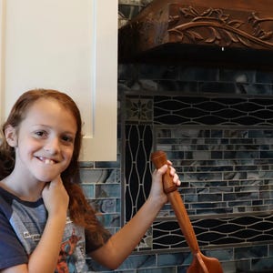 May include: A wooden meat chopper is held over a black skillet with ground meat. The kitchen features a decorative backsplash and a wooden range hood with floral carvings. A young person is smiling in the foreground.