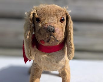 以下が含まれることがあります： 茶色の毛並みと垂れ耳が特徴的な、ヴィンテージの犬のぬいぐるみ。犬は暗い色の目を持ち、首には赤いリボンが巻かれています。おもちゃは白い表面に立っており、背景はぼやけた木材です。