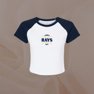 May include: White and navy blue baseball-style shirt with the text "TAMPA RAYS BAY" in navy blue and yellow. The shirt has navy blue sleeves and a white body. The text is centered on the chest.