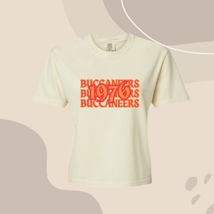 May include: A white t-shirt with a graphic design featuring the text "BUCCANEERS 1976 BUCCANEERS" in orange and red.