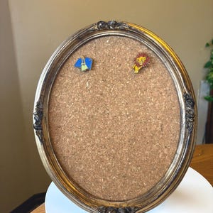 May include: Oval-shaped corkboard with a gold-colored ornate frame. Two enamel pins are attached to the corkboard: one depicting a blue and green object, and the other a yellow lion with a red mane. The corkboard is displayed on a white surface.