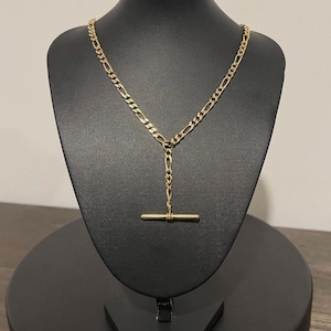 May include: A gold-toned necklace featuring a figaro chain design that transitions into a Y-shaped drop. The necklace terminates in a horizontal bar toggle clasp, which is suspended from a central vertical chain segment, creating a clean aesthetic.